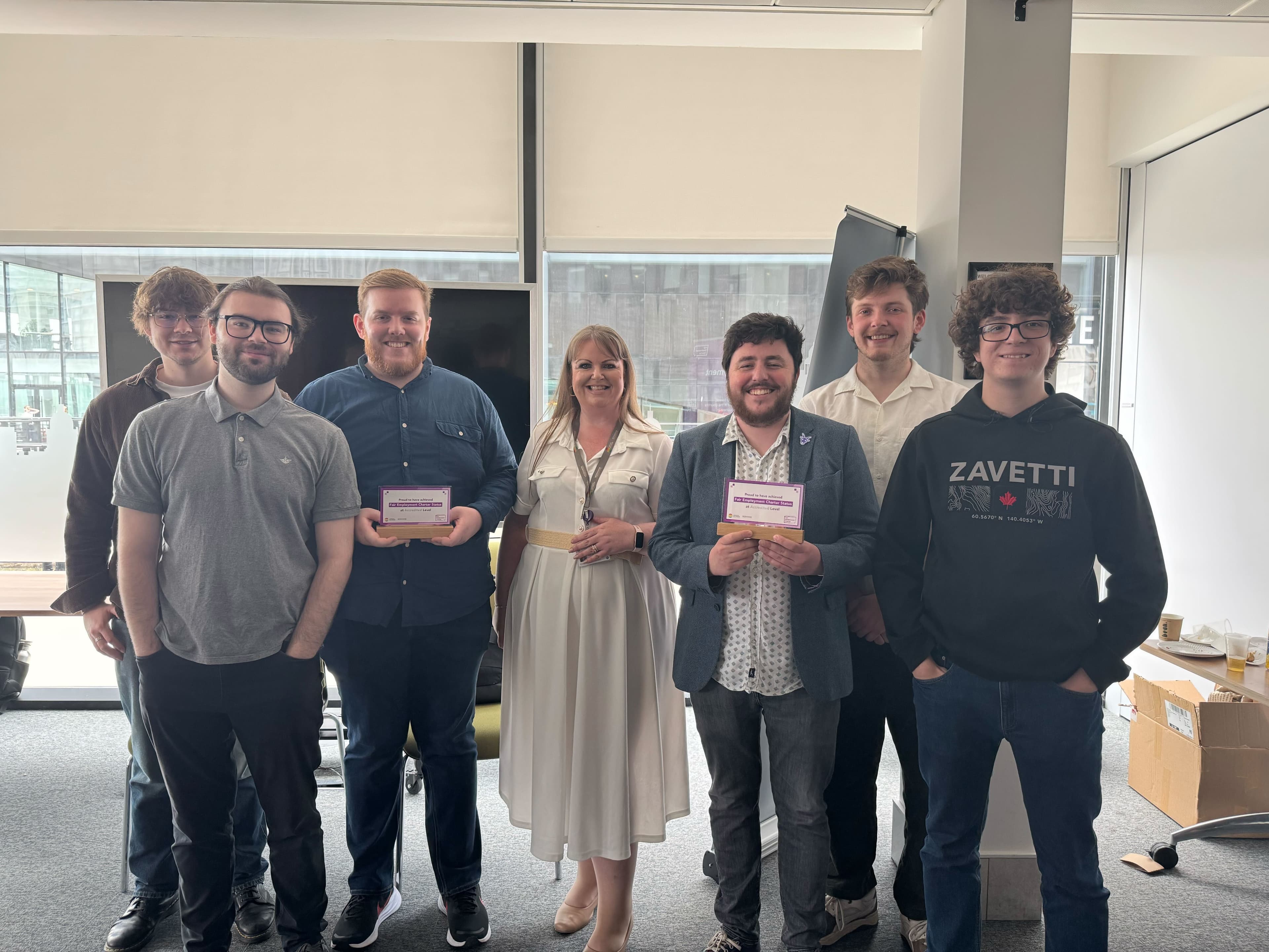 A group of Jamescape and Harker staff stood together in a conference room, smiling towards the camera, alongside LCRCA staff. Two staff are holding Fair Employment Charter Accredited plaques.