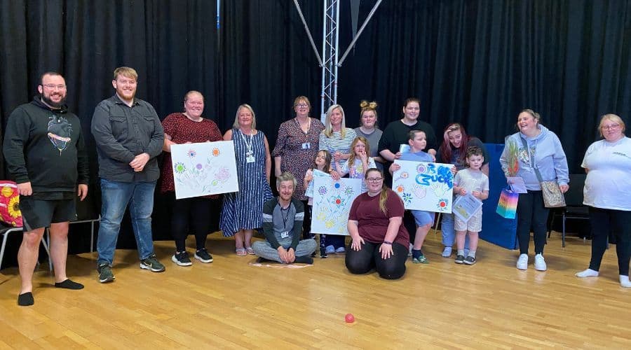 A group of parents, children, and support workers stood together smiling towards the camera in a theatre hall.