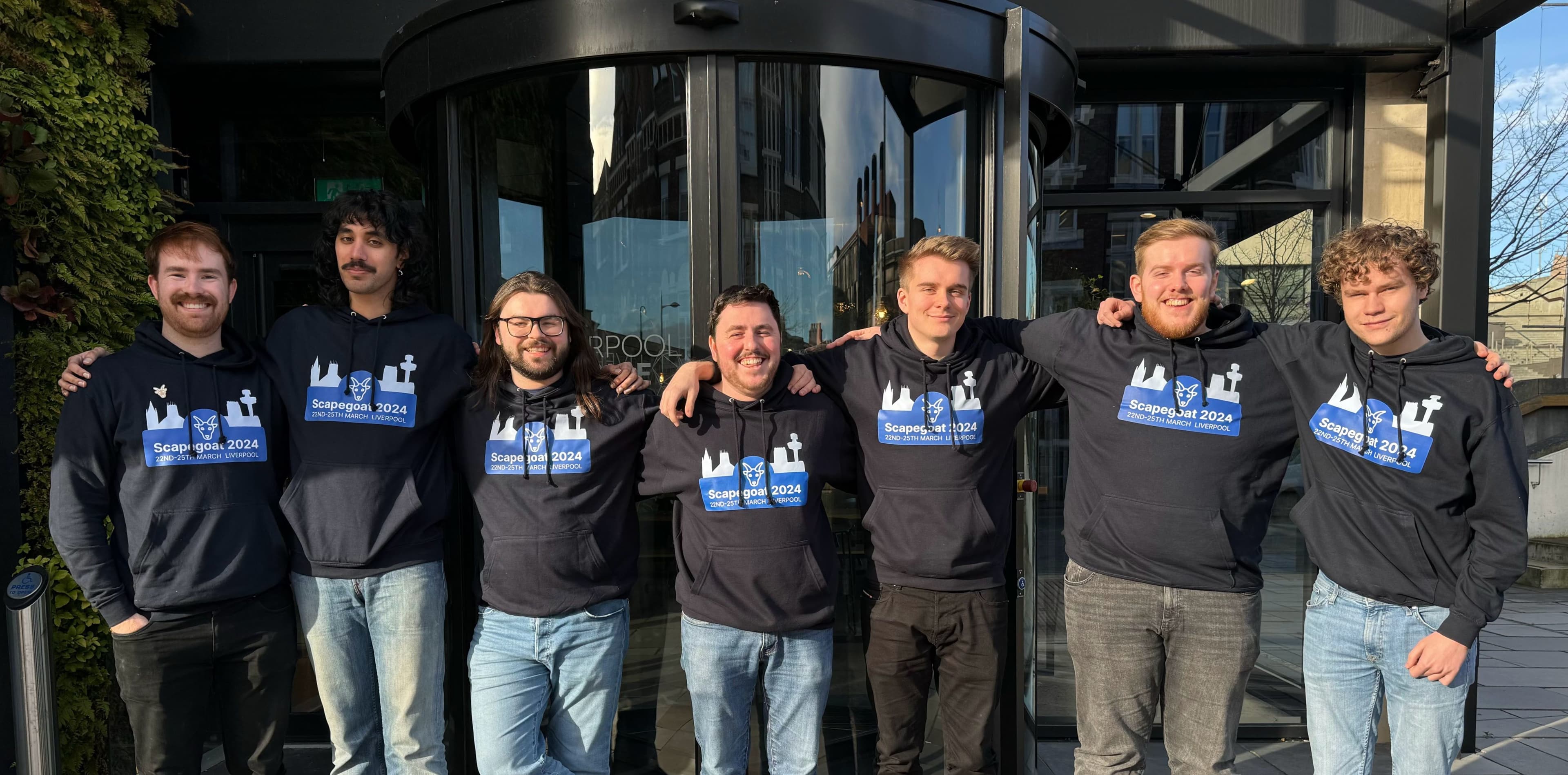 Jamescape staff outside the Liverpool Science Park entrance with their arms around each other's shoulders. All staff are wearing Scapegoat branded hoodies.